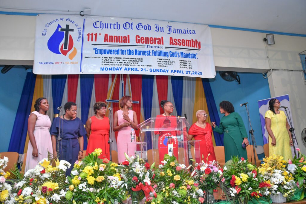 A group of women standing on a stage during the 111th Annual General Assembly of the Church of God in Jamaica, with a colorful banner in the background and flowers in the foreground.