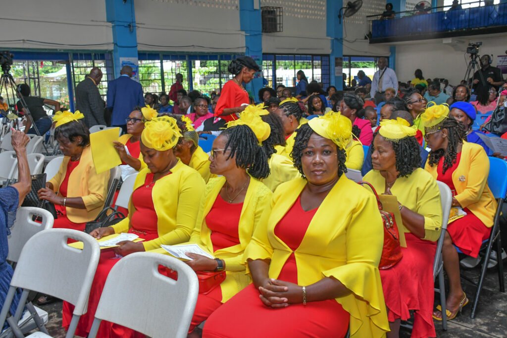 A group of women wearing yellow and red outfits, some with matching hats, seated in an auditorium filled with attendees.