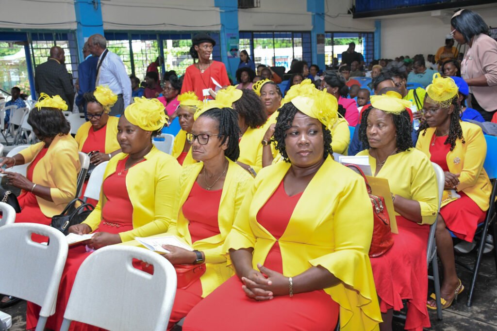 A group of women dressed in bright yellow jackets and red dresses, wearing matching yellow hats, seated in a hall filled with an audience.