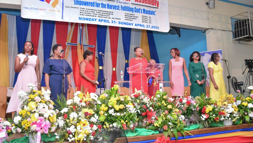 A group of women speaking at a podium during an assembly event, adorned with colorful flowers and decorations, with a banner in the background detailing the event theme and dates.