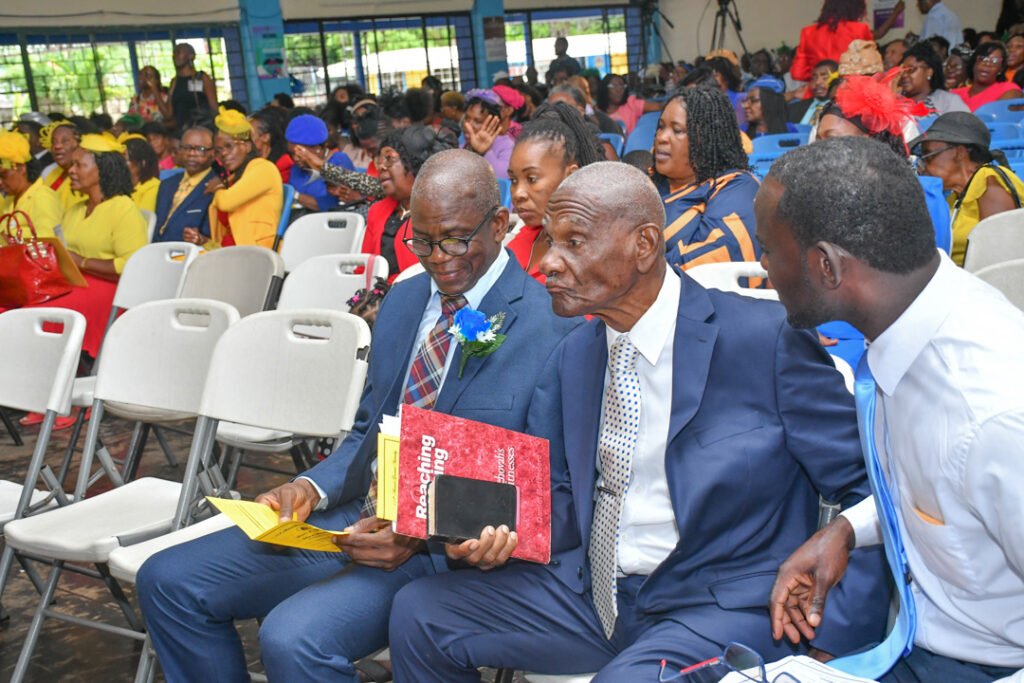 A group of people sitting in a hall during an event, with attendees wearing colorful outfits. Two men in formal attire are engaged in conversation, one holding a program or booklet. The background features a diverse audience, mostly women, in various colorful attire.
