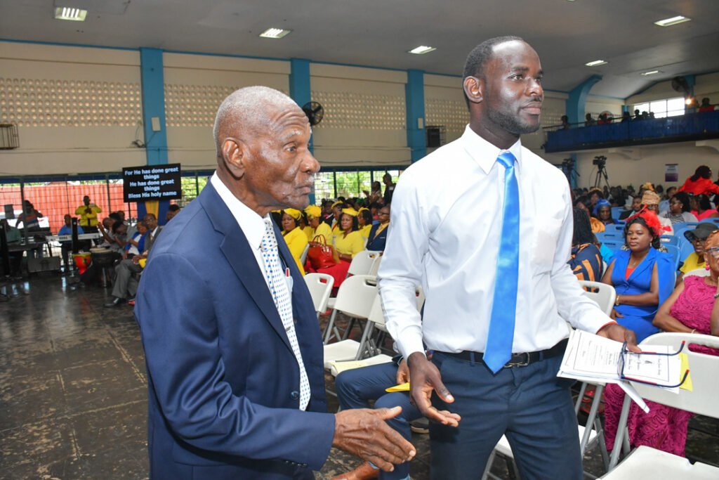 An elderly man in a blue suit and tie interacts with a younger man in a white shirt and blue tie at a gathering. In the background, an audience of people dressed in colorful outfits watches the event.