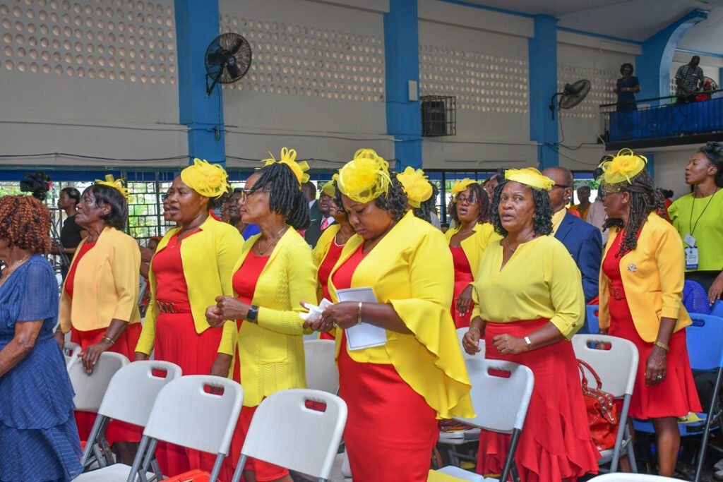 A group of women dressed in red and yellow outfits, singing passionately in a gathering, with some wearing matching hats. The setting appears to be a spacious indoor venue with seating and a blue accent wall.