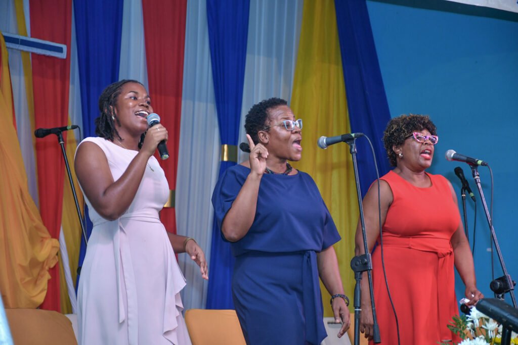 Three women singing on stage with microphones, dressed in colorful outfits, in front of a decorated backdrop.