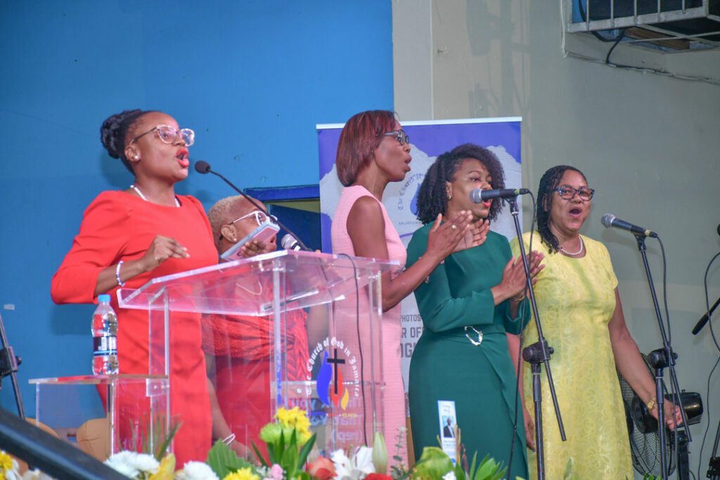 A group of four women singing passionately at a church event, standing behind a transparent podium with flowers in the foreground.