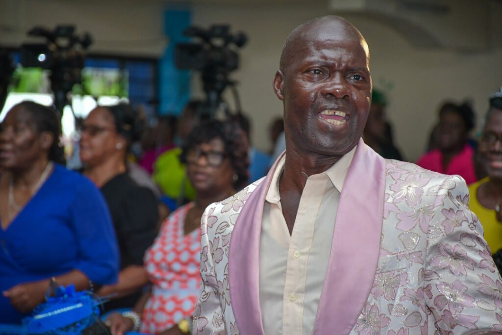 A man in a light-colored suit with floral embellishments smiles and speaks to a crowd, while various attendees in the background appear engaged and dressed in colorful clothing.