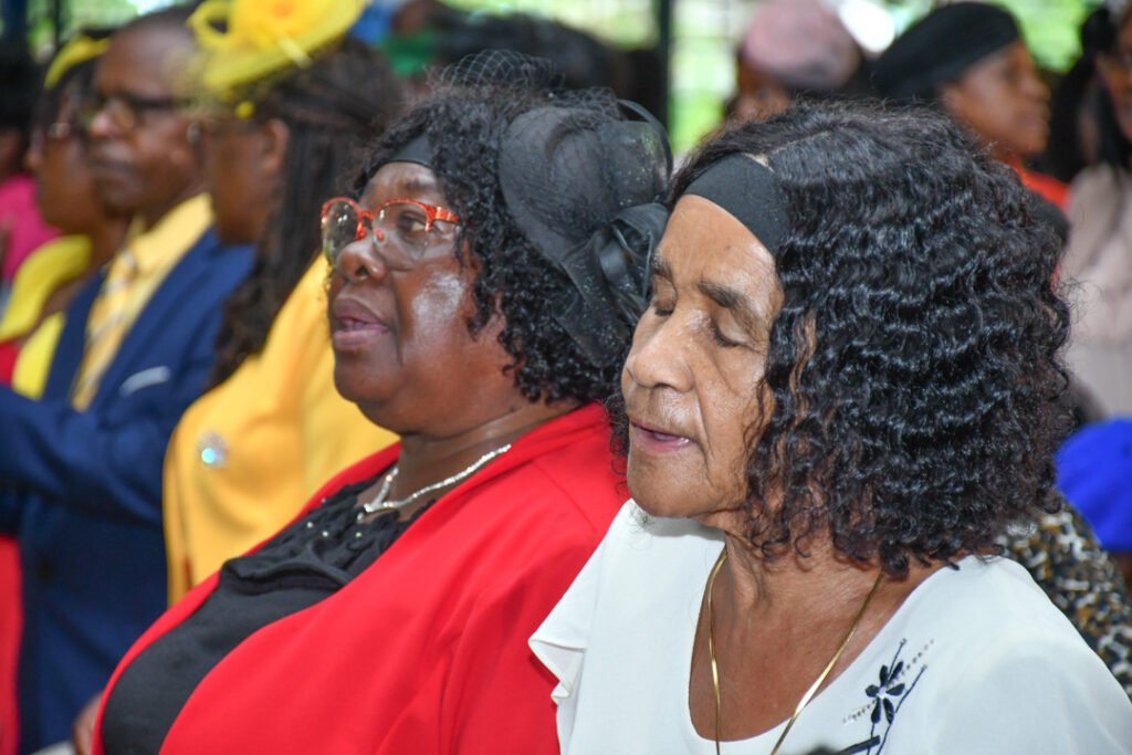 Two women with curly hair attending a gathering, wearing colorful attire and looking contemplative. A group of people is visible in the background.