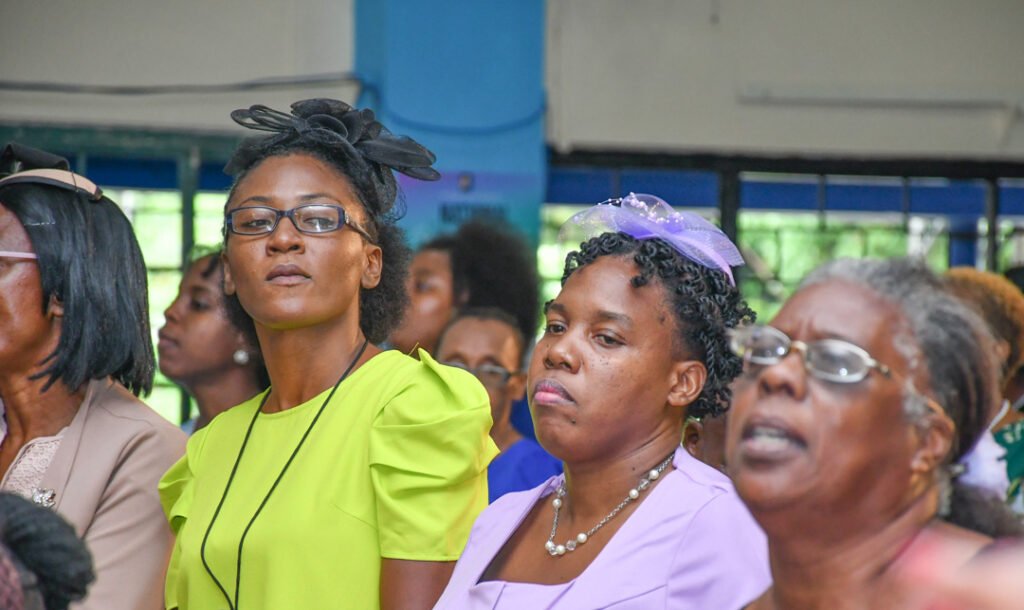 A group of women attending an event, some wearing colorful dresses and hats, with expressions of engagement.