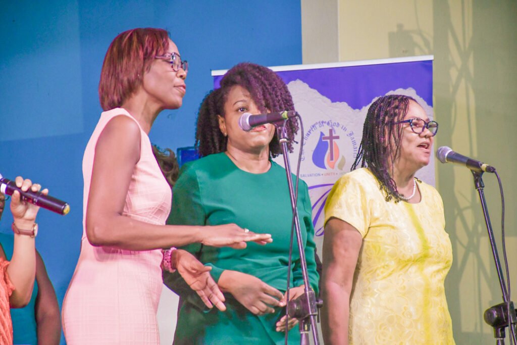 Three women singing together on stage, holding microphones and wearing colorful dresses, with a backdrop featuring a religious emblem.