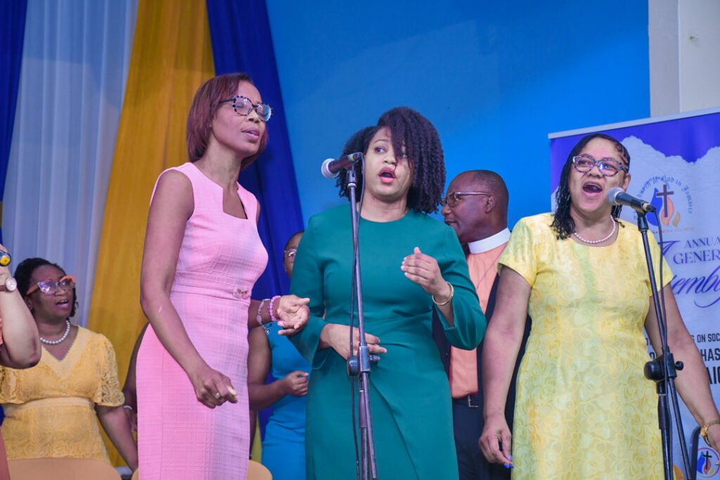 A group of people singing at an event, with a focus on three women in colorful outfits standing at the front. The backdrop features draped fabric in yellow and blue, and a banner on the side.