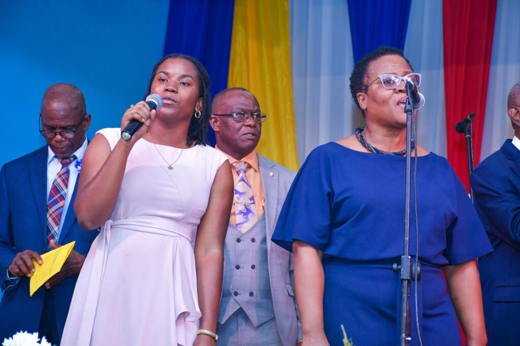 A group of individuals performing on stage, with a female singer holding a microphone in the foreground, while several others stand behind her, some wearing formal attire. The background features colorful drapes.