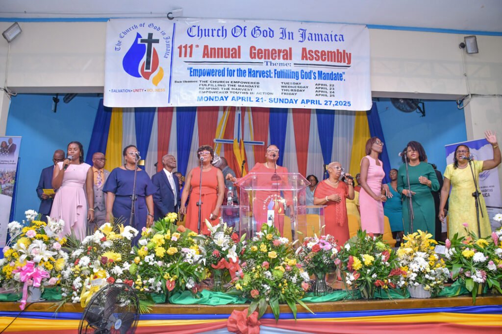 A group of singers performing at the 111th Annual General Assembly of the Church of God in Jamaica, with colorful floral arrangements and a banner displaying the event details in the background.