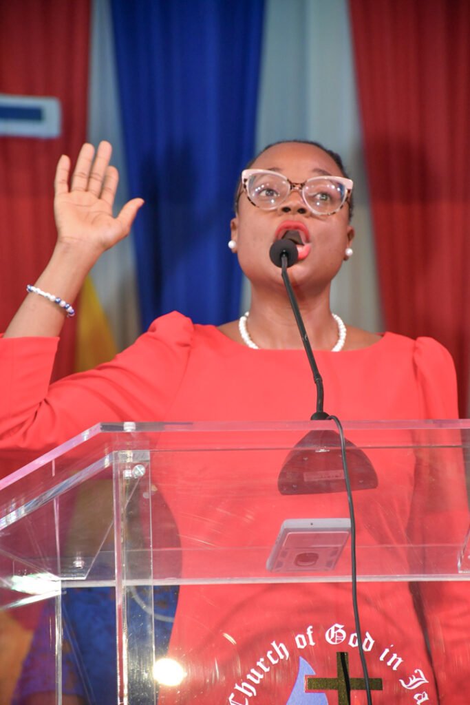 A woman in a red dress stands at a clear podium, passionately speaking into a microphone, with her right hand raised. Behind her are colorful drapes and the logo of the Church of God in Jamaica.