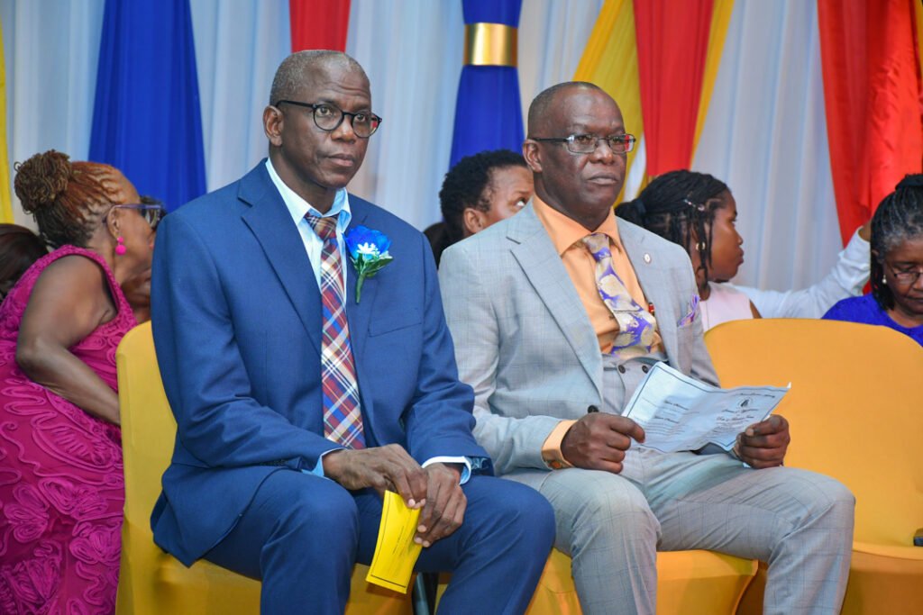 Two men seated in formal attire, one wearing a blue suit with a plaid tie and the other in a gray suit with an orange shirt. They are at an event with colorful drapes in the background.