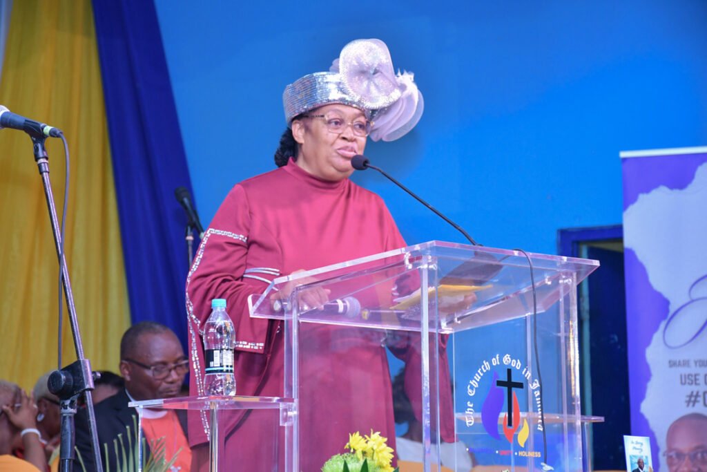 A woman in a burgundy dress and a decorative hat speaks at a podium during an event at a church, with attendees in the background.