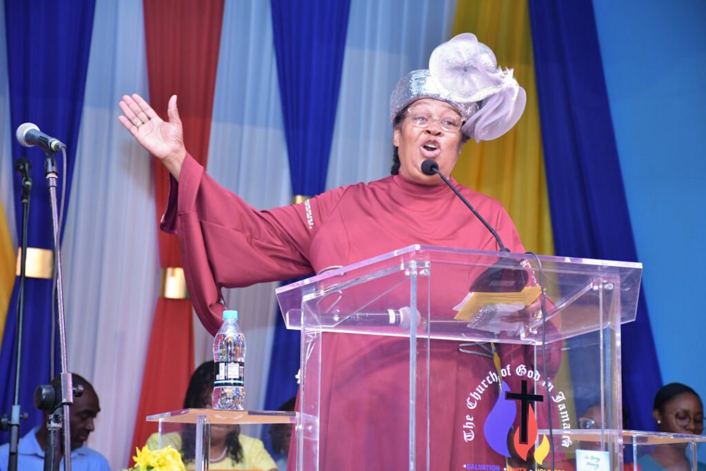 A speaker passionately delivering a message at a podium with a microphone, dressed in a red garment and a decorative hat, in front of a colorful backdrop.