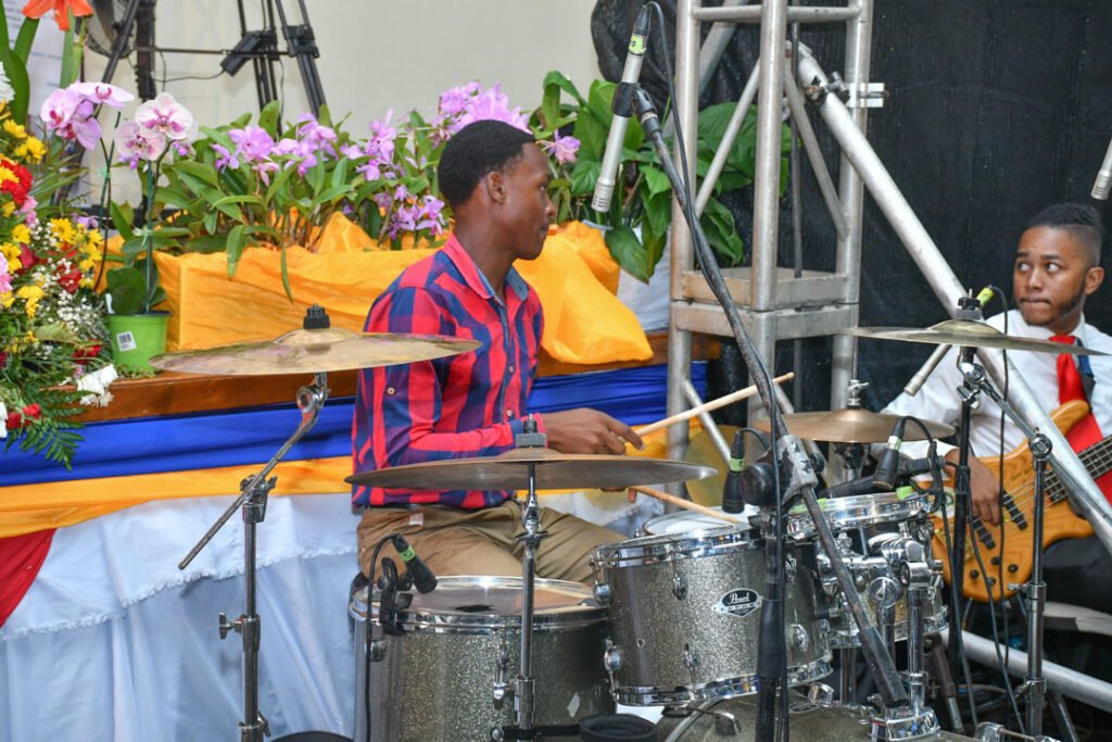 A drummer playing a silver drum set in front of a vibrant flower arrangement, with a bassist seated nearby.