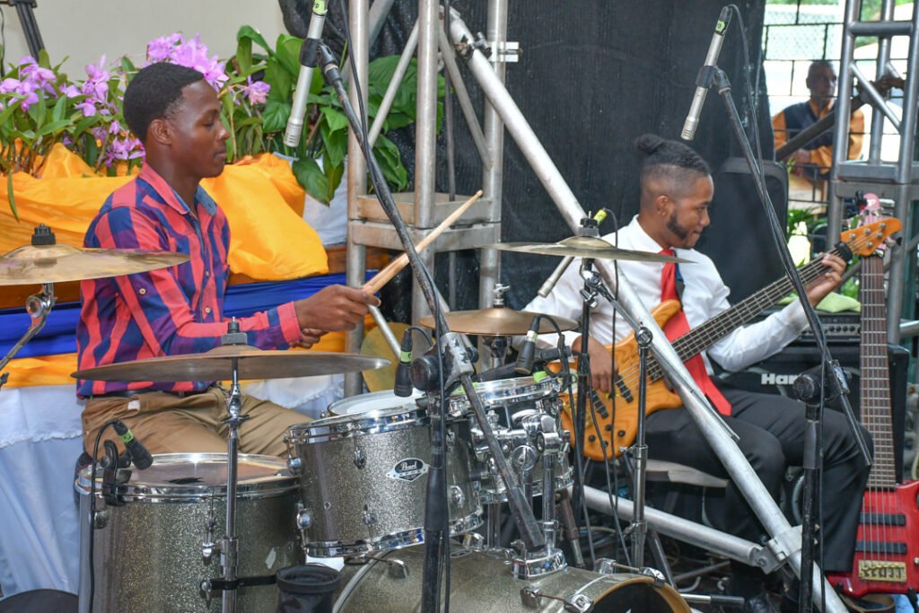 A drummer playing on a silver drum set and a bassist playing an electric guitar during a performance, surrounded by purple flowers and stage equipment.