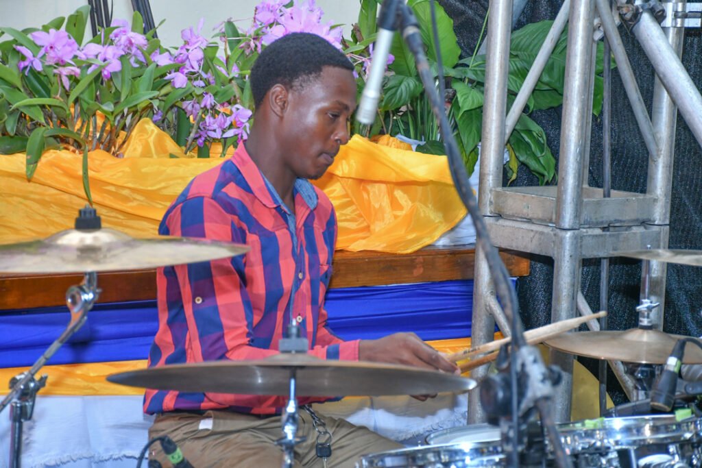 A young man playing the drums on stage, surrounded by potted plants and colorful fabric decorations.