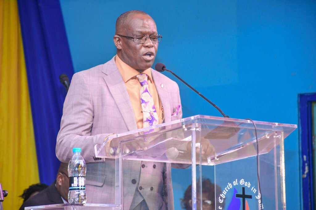 A man in a light purple suit speaks passionately at a podium during a church event, with a blue background and decorative fabrics in view.