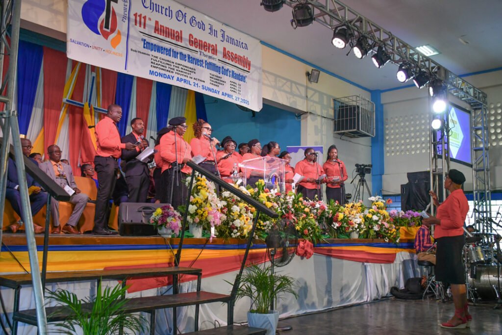 A choir dressed in orange performs on stage during the 111th Annual General Assembly of the Church of God in Jamaica, with a colorful floral arrangement in the foreground.