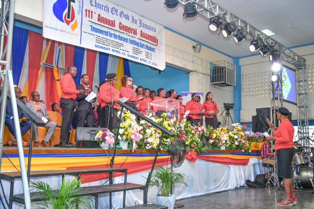 A choir dressed in red uniforms performs on stage at the Church of God in Jamaica's 111th Annual General Assembly, surrounded by colorful flowers and decorations.