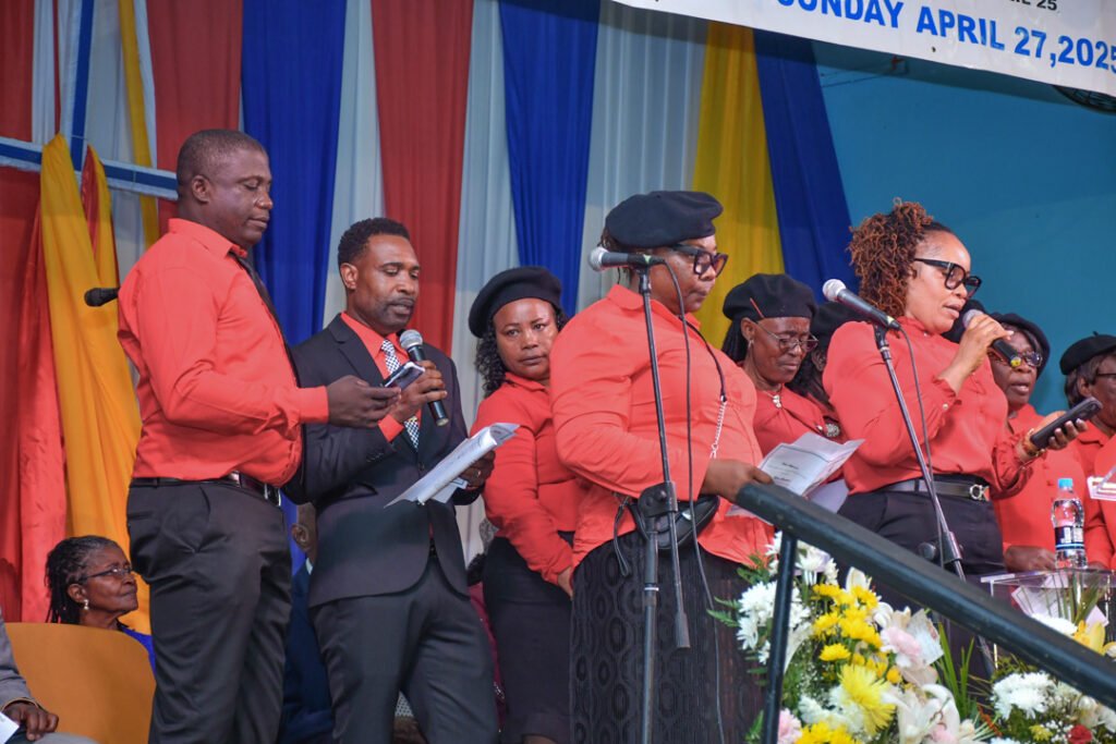 A group of people in red shirts and black hats performing at a podium, with a colorful backdrop and floral arrangements.