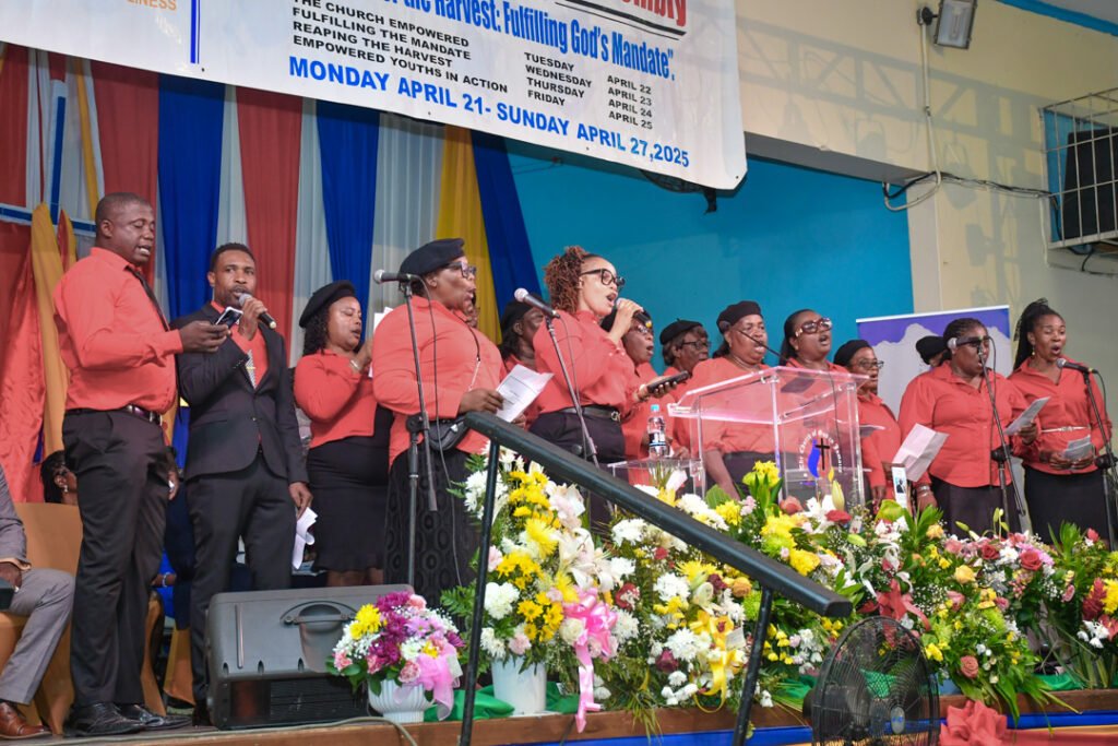 A choir singing on stage, wearing matching red shirts, with a microphone and a transparent podium in front, surrounded by colorful flowers and banners in the background.