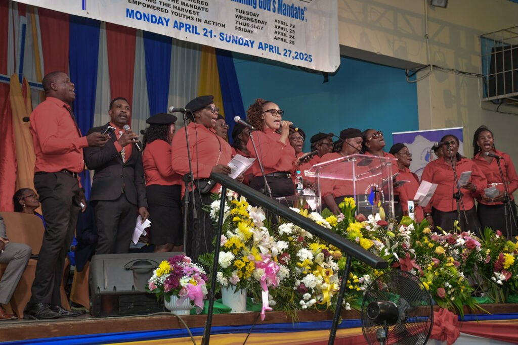 A choir dressed in red and black performs on stage surrounded by colorful floral arrangements and banners, with a vibrant atmosphere.