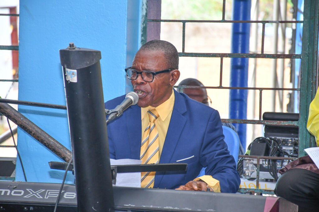 An elderly man wearing glasses and a blue suit is singing into a microphone while playing a keyboard. The setting appears to be a performance space with a blue wall in the background.