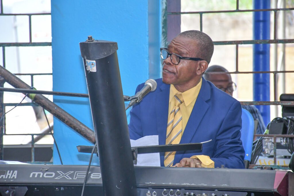 A man in a blue suit and yellow shirt playing a keyboard and singing into a microphone at a performance or event.