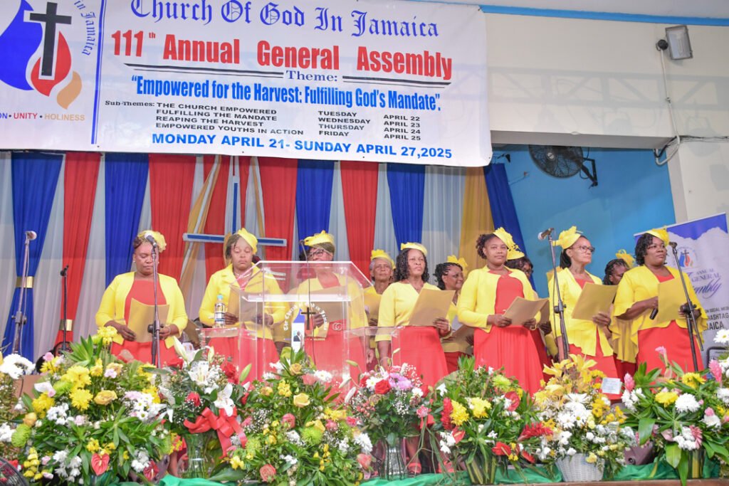 A choir dressed in vibrant yellow and red outfits performs during the 111th Annual General Assembly of the Church of God in Jamaica, with a colorful backdrop and floral arrangements.