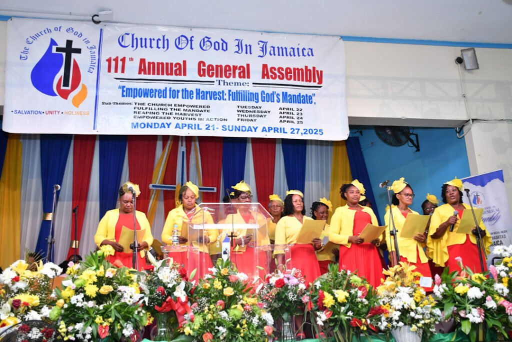 A group of women in yellow and red attire perform during the 111th Annual General Assembly of the Church of God in Jamaica, with a banner in the background displaying the event's theme and details.