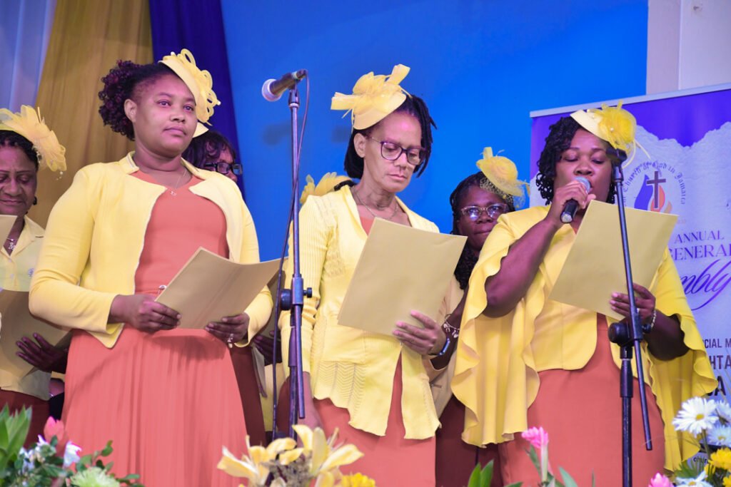 A group of women wearing yellow outfits and hats, singing and holding papers during an event.