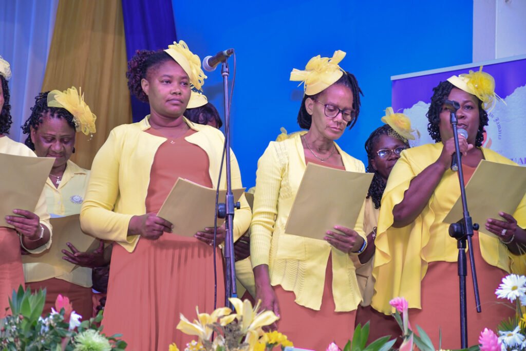 A group of women dressed in yellow outfits, some wearing matching hats, standing together and holding sheets of paper while singing at an event.