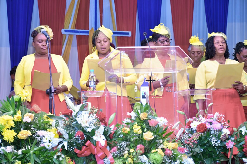 Group of women singing at a church service, wearing matching yellow and orange outfits, with floral decorations in the foreground.