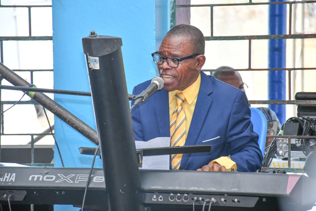A man in a blue suit and yellow shirt plays a keyboard while singing into a microphone, with a blue wall and equipment visible in the background.