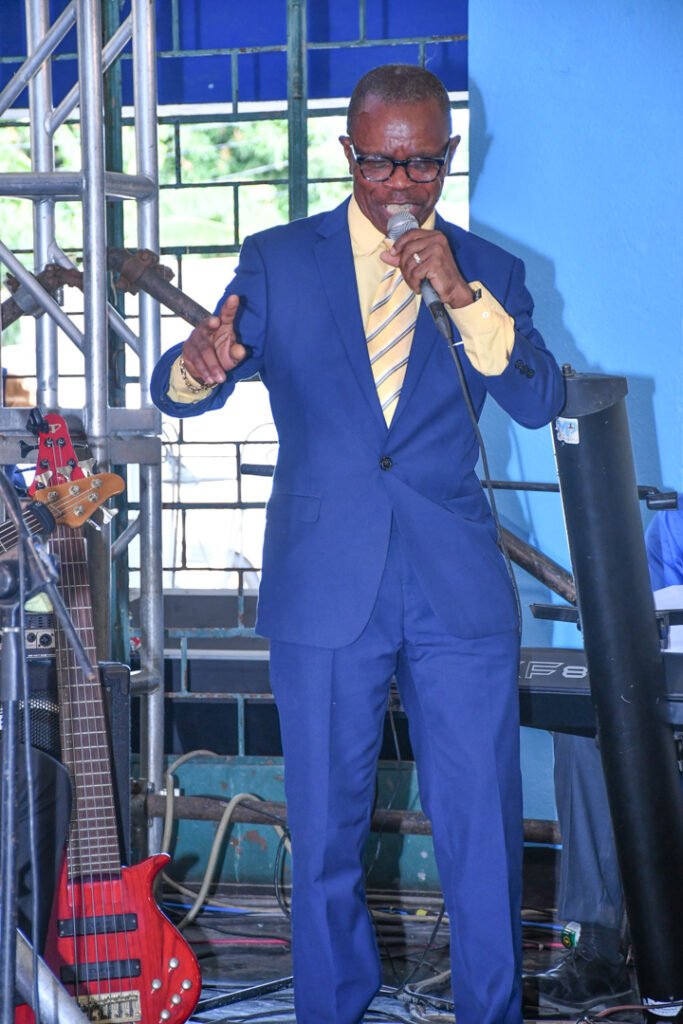 A man in a blue suit and yellow tie speaking into a microphone on stage, with a red bass guitar nearby.