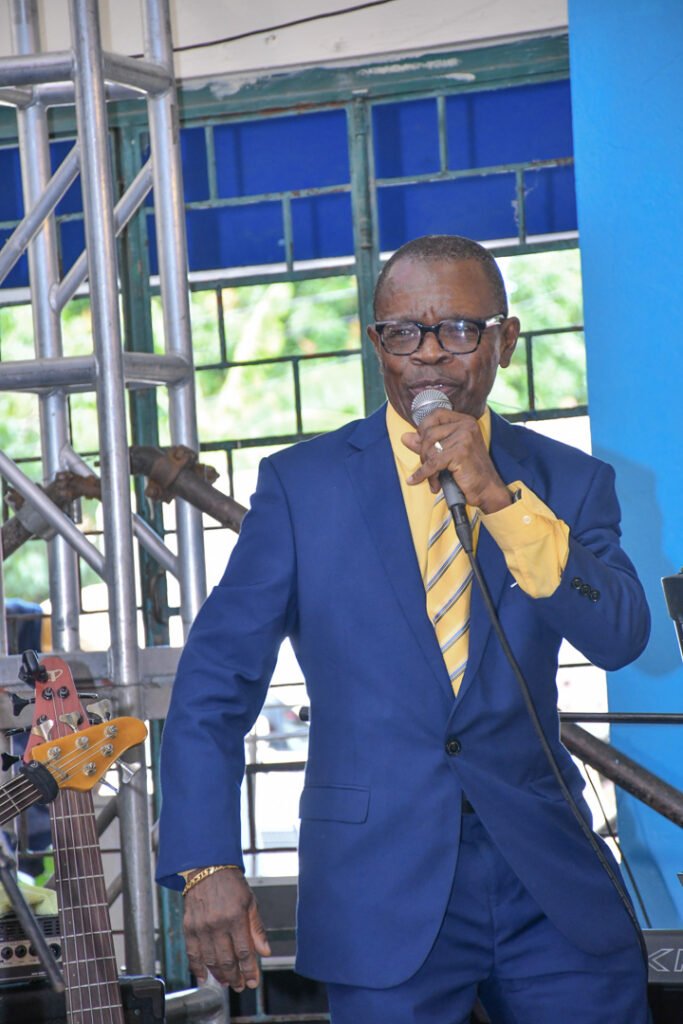 An older man in a blue suit and yellow shirt is speaking into a microphone, performing in front of a blue backdrop. A bass guitar is partially visible in the foreground.