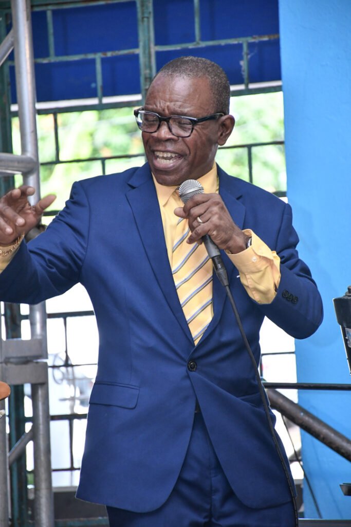 A man in a blue suit and yellow shirt speaks passionately into a microphone, gesturing with his hands, against a backdrop of blue and green.