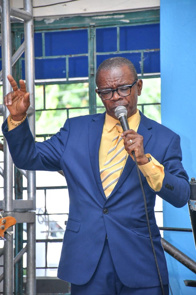 A man in a blue suit with a yellow shirt and striped tie speaks into a microphone, gesturing with one hand, against a backdrop of blue walls and windows.