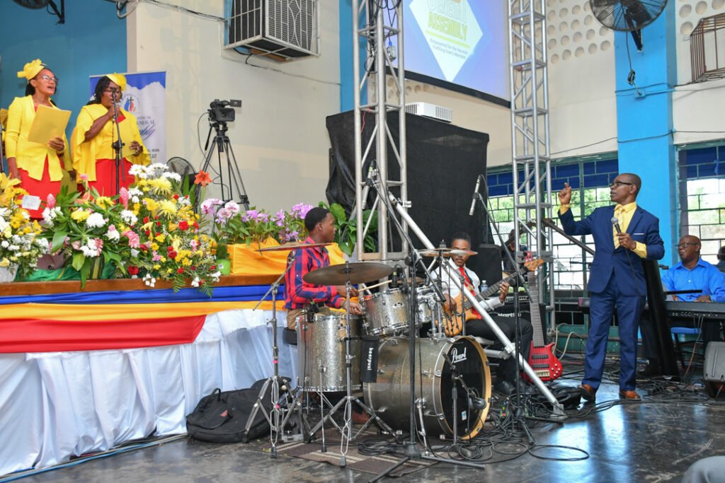 A group of singers in bright yellow and red attire performing on stage, accompanied by a band. The scene is decorated with colorful flowers on the stage, and a speaker is seen addressing the audience.
