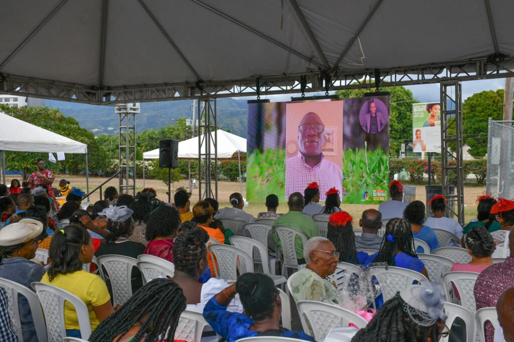 A large outdoor gathering with a diverse audience seated in white chairs under a tent. A big screen displays a speaker giving a presentation, while attendees engage with the event.