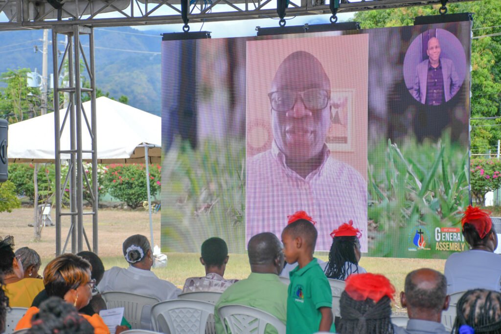 A large screen displaying a video call of a man wearing glasses, speaking during an event, with an audience seated in front of it.