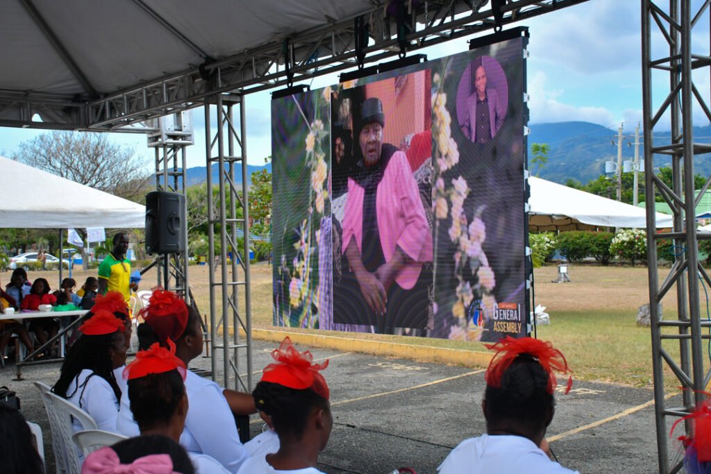 A large screen displaying a speaker during an outdoor event, with attendees sitting in front wearing matching outfits and red accessories.