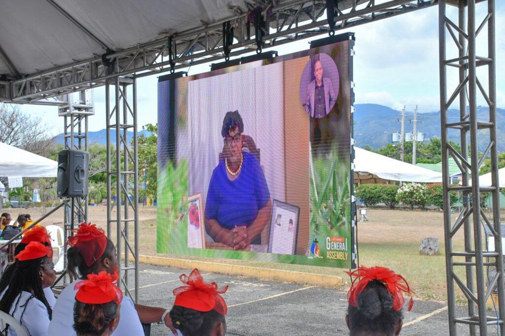 A large screen displays a virtual meeting with a woman in a blue outfit and pearls sitting in front of a framed picture. Outdoor audience members with red bows in their hair are seated in front.