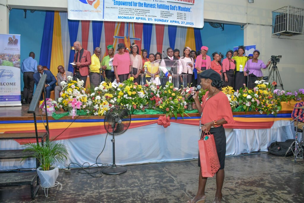 A group of people in colorful clothing gathered on stage at a church event, with a flower display in front and a woman in a red top speaking into a microphone.