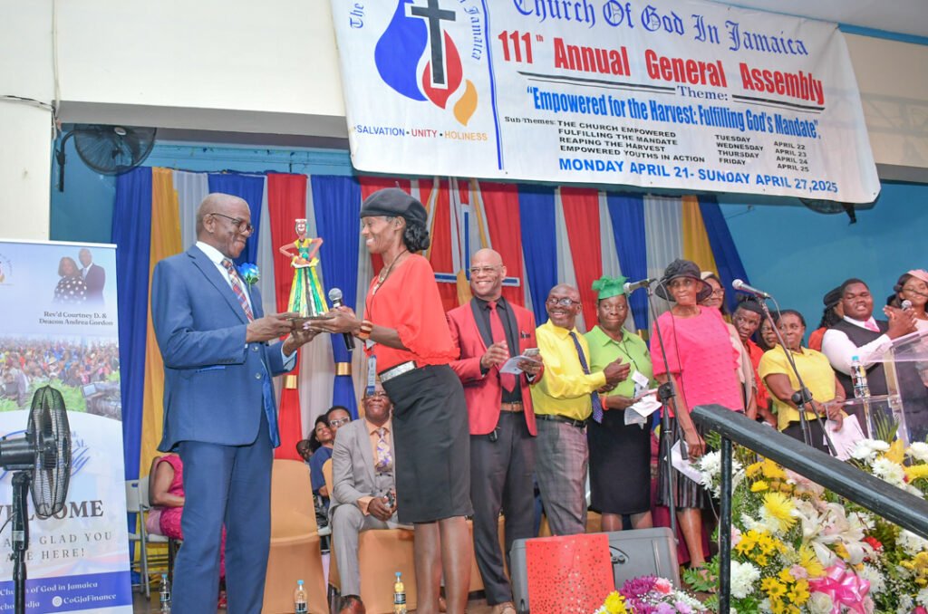 A group of people at a church event in Jamaica, with one person presenting a decorative doll to another. The background features a colorful banner and various attendees in different outfits.