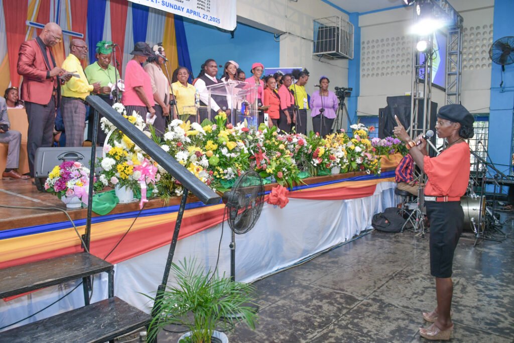 A group of singers performing on stage at a colorful event, surrounded by flower arrangements and a podium, with a woman in the foreground gesturing as she addresses the audience.