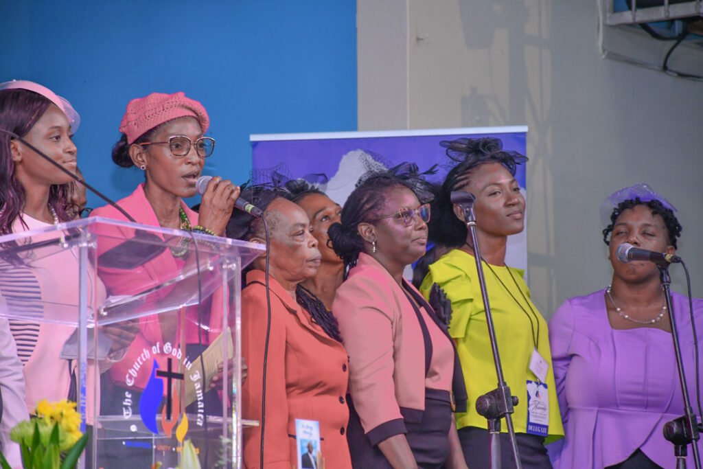 A group of women singing on stage during a church event, with one woman speaking into a microphone. They are dressed in colorful outfits, with a backdrop featuring a church logo.